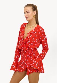 Vestido floral rojo con mangas largas, escote con volantes y cintura ceñida por un cinturón. Presenta patrones de flores rosas y puños acampanados. Tela ligera.