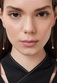 Silverfärgade kedjeörhängen med cirkulära detaljer, som har en slät yta och minimalistisk design. Modellen bär en svart topp med vit sömnad.