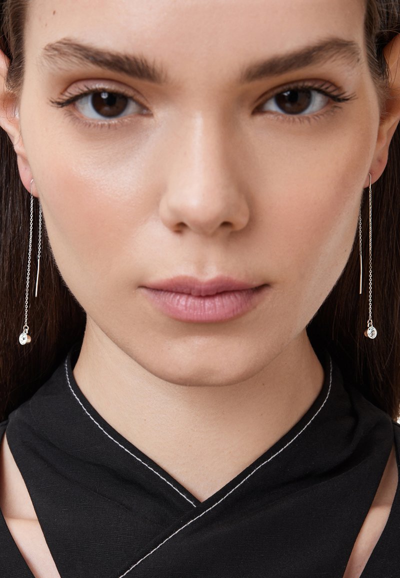 Silverfärgade kedjeörhängen med cirkulära detaljer, som har en slät yta och minimalistisk design. Modellen bär en svart topp med vit sömnad.