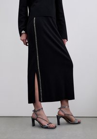 Mujer con falda larga negra con abertura lateral y detalle de franja plateada, combinada con sandalias de tacón alto plateadas con tiras, sobre suelo gris.