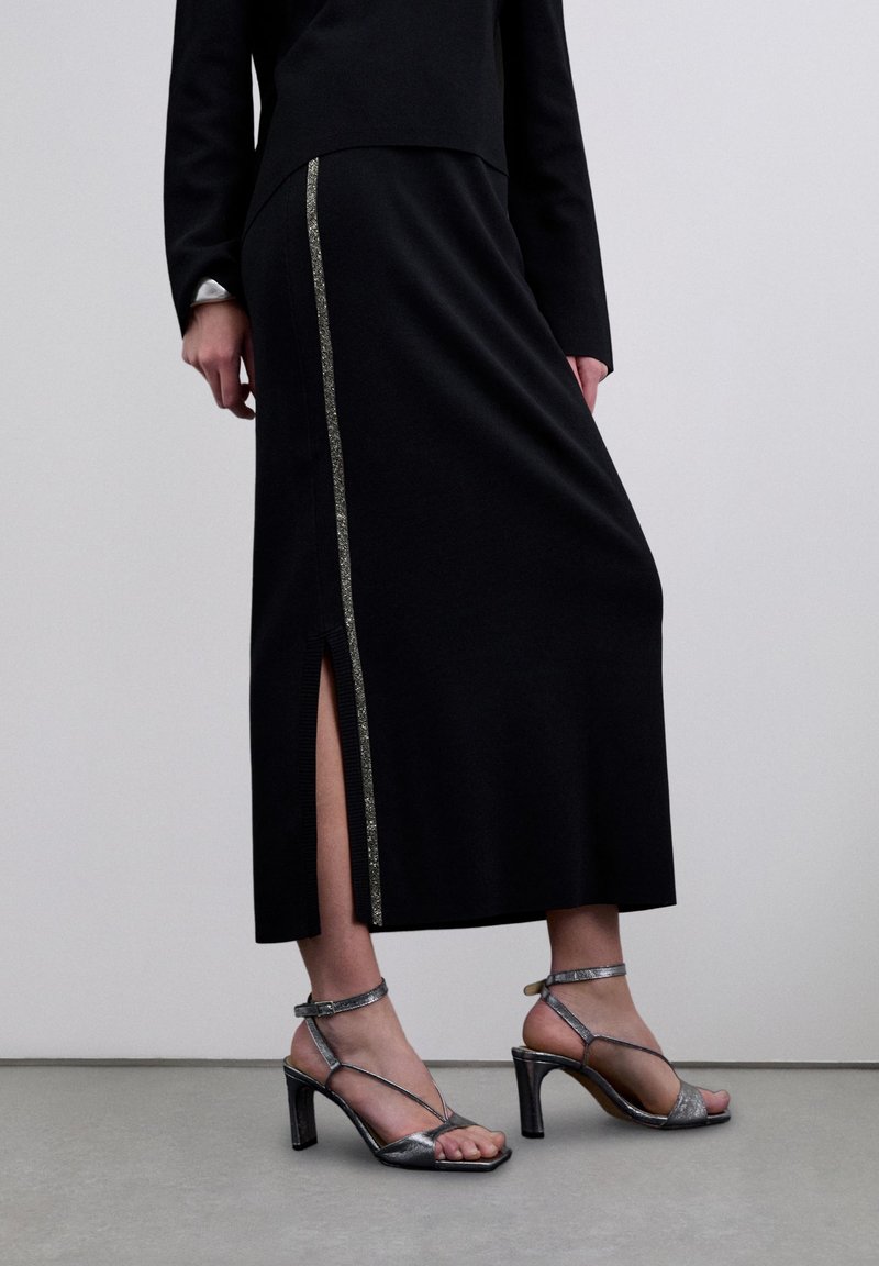 Mujer con falda larga negra con abertura lateral y detalle de franja plateada, combinada con sandalias de tacón alto plateadas con tiras, sobre suelo gris.