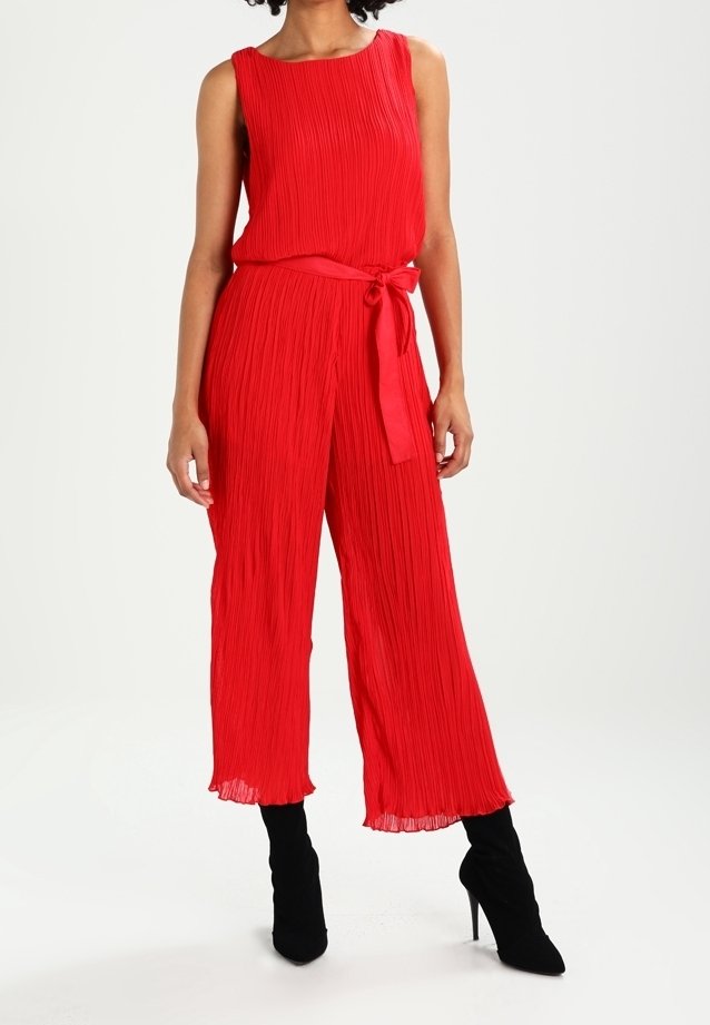 Femme portant une combinaison plissée rouge sans manches avec une ceinture nouée à la taille et des bottines à talons hauts noires, se tenant devant un fond blanc.