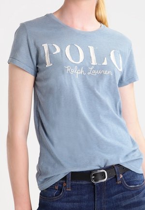 Kobieta nosząca jasnoniebieską koszulkę Polo Ralph Lauren wpuszczoną w ciemnoniebieskie dżinsy z czarnym paskiem i srebrną klamrą.