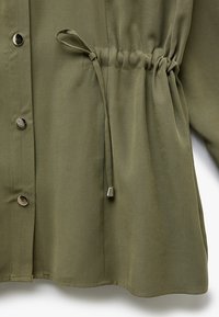 Camicia verde oliva in tessuto testurizzato, con bottoni a scatto argentati e vita con cordino per una vestibilità regolabile.