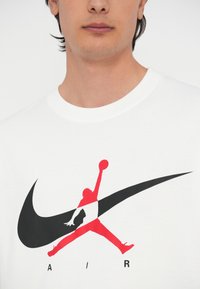 Λευκό μπλουζάκι με μεγάλο μαύρο λογότυπο Nike και κόκκινη σιλουέτα Jumpman με τη λέξη "AIR" τυπωμένη από κάτω σε μαύρο. Λείο βαμβακερό υλικό.