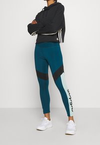 Czarna krótkia bluza z kapturem w białe paski i logo, sparowana z legginsami w kolorze teal i czerni z blokami kolorów, z białym akcentem logo. Białe sneakersy.