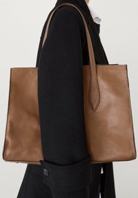 Sac tote en cuir marron avec une texture lisse, une forme rectangulaire et de longues poignées. Détails cousus le long des bords, tenu contre un manteau noir.