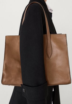 Borsa tote in pelle marrone con una texture liscia, forma rettangolare e manici lunghi. Dettagli cuciti lungo i bordi, appoggiata su un cappotto nero.