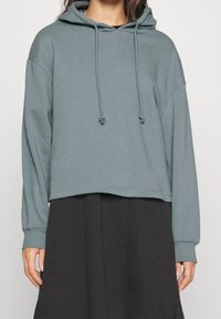 Sudadera recortada de color azul claro con cordones, de tejido suave, puños acanalados y silueta relajada, llevada sobre una prenda negra.