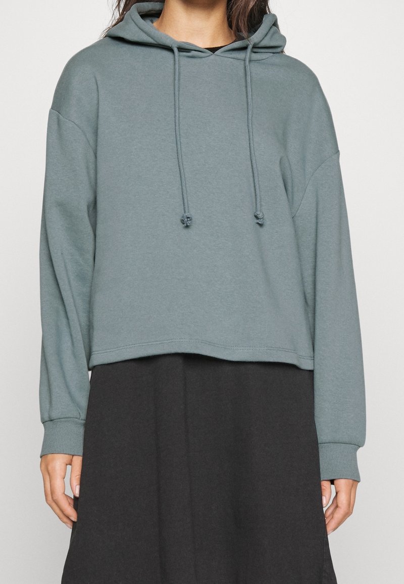 Sudadera recortada de color azul claro con cordones, de tejido suave, puños acanalados y silueta relajada, llevada sobre una prenda negra.