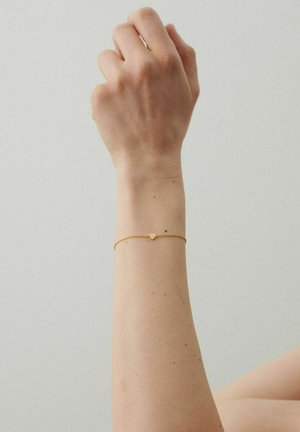Løftet underarm med et delikat armbånd af guld med en lille hjertevedhæng mod en ensfarvet lys baggrund.