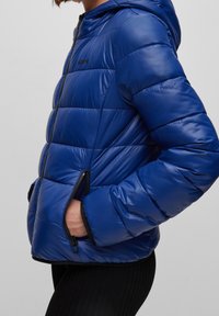 Blå pufferjacka med huva, med en blank yta, horisontell sömnad och sidofickor med dragkedja. Svarta detaljer på ärmar och nederkant.