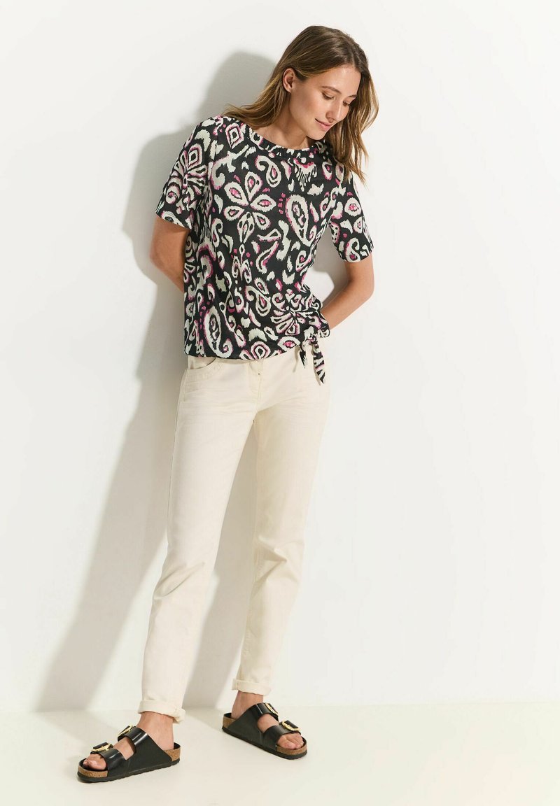 Top negro de manga corta con un patrón floral colorido, combinado con unos pantalones vaqueros color crema y sandalias negras con hebillas doradas.