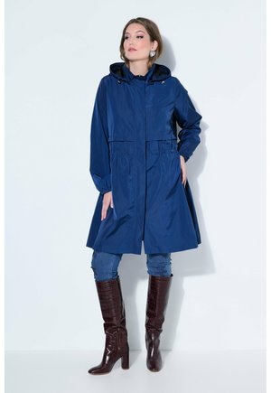 Femme portant un manteau à capuche bleu, un jean bleu, des bottes montantes marron et de grandes boucles d'oreilles argentées, debout devant un fond blanc.