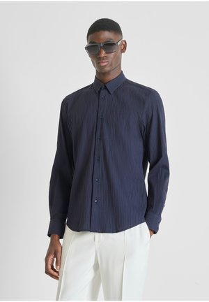 Man draagt donkerblauw, gestructureerd overhemd met lange mouwen en knopen, witte broek en grote zwarte zonnebril tegen een effen grijze achtergrond.