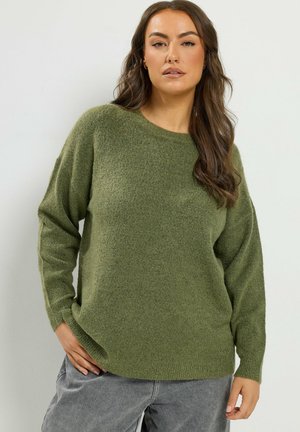 Femme aux longs cheveux bruns portant un pull en tricot vert et un pantalon gris, debout devant un fond clair uni.