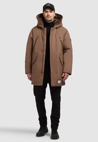 Braune gepolsterte Parka mit abnehmbarer Kapuze, vorne mit Reißverschluss und Druckknopfverschlüssen, zwei seitlichen Taschen und schwarzen Akzenten. Über einem schwarzen Rollkragenpullover getragen.