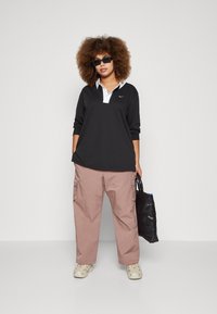 Polo noir à manches longues avec col blanc et logo Nike, associé à un pantalon cargo rose clair. Porte des lunettes de soleil et tient un sac noir.