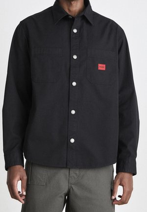 Chemise noire à boutons avec deux poches poitrine, dont une ornée d'une petite étiquette rouge "HUGO", portée avec un pantalon gris.