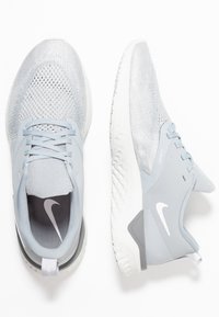 Zapatillas Nike de color gris claro con parte superior de malla texturizada, acentos lisos contrastantes y una suela de goma con un patrón ondulado.