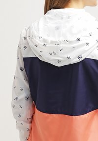 Farvet blokket jakke med en hvid hætte med anker- og kompasmønstre, en mørkeblå midtersektion og en koral nederste sektion. Glat stof.