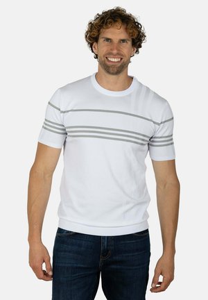 Hombre sonriente con cabello rizado que lleva una camisa blanca de manga corta con rayas horizontales grises y vaqueros azul oscuro sobre un fondo liso.