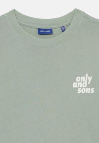 Svetlozeleni bombažni majica s kratkimi rokavi in krojnim ovratnikom, na levi strani ima majhen bel logotip z napisom "only and sons". Gladka tekstura.