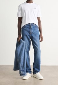 Wit katoenen T-shirt met "Hugo"-logo, blauwe spijkerbroek en een blauwe spijkerjasje in de hand. Witte sneakers, ontspannen pasvorm, gladde textuur.