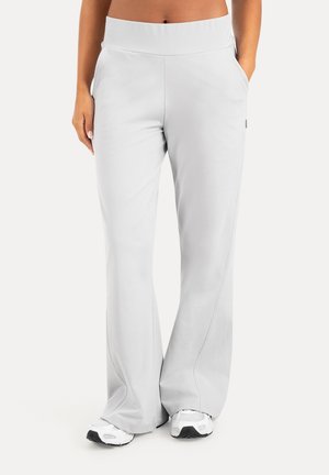 Mujer con pantalones anchos gris claro con bolsillos laterales y zapatillas blancas, de pie con una mano en el bolsillo.