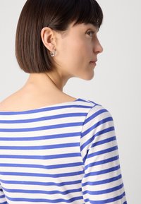 Stripetopp i hvit og blå, med båtutringning og trekvartlange ermer. Sølvøredobber pryder modellens ører.