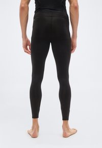 Άνδρας που στέκεται ξυπόλητος, με την πλάτη γυρ ισμένη, φορώντας εφαρμοστά μαύρα αθλητικά κολάν και μαύρη αμάνικη μπλούζα, σε απλό φωτεινό φόντο.