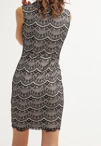 Svart spetsklänning med ärmlös design, prydd med ett vågigt mönster och intrikata blommor. Rundad nederkant med spetsdetalj.