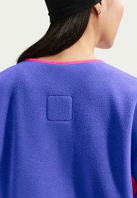 Γυναίκα που φοράει ένα μοβ fleece ρούχο με ανάγλυφο τετράγωνο σχέδιο στην πλάτη και ροζ μπορντούρα κατά μήκος της λαιμόκοψης, μαύρο καπέλο και χρυσό σκουλαρίκι κρίκο.