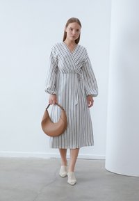 Gestreiftes, langärmliges Wickelkleid mit einem Bindegurt, das helle und dunkle Streifen aufweist. Das Model hält eine braune, geschwungene Ledertasche.