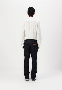 Donkerblauwe spijkerbroek met zichtbare stiksels en een leren patch op de rug; gecombineerd met een witte blouse met lange mouwen, gezien vanuit de achterkant.