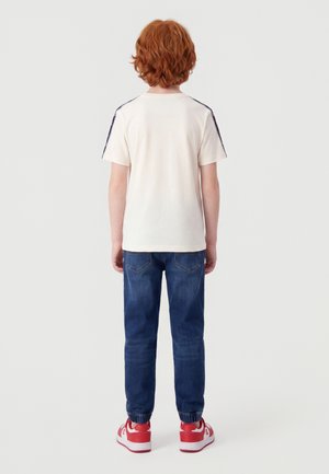 Rude dziecko stoi tyłem, ubrane w biały T-shirt w niebieskie paski, niebieskie dżinsy oraz czerwono-białe buty sportowe.