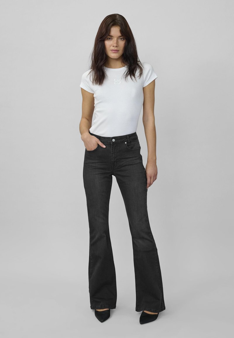 Femeie cu păr închis la culoare, purtând un tricou alb mulat, blugi negri evazați cu talie înaltă și pantofi negri cu vârful ascuțit, stând cu o mână în buzunar.