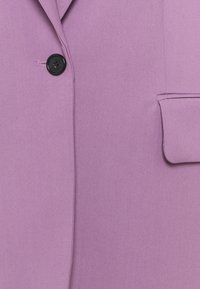 Blazer lavande avec un bouton noir fixé près du col et une poche à rabat sur le côté droit.