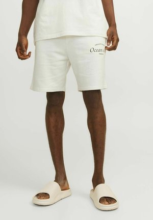 Shorts en coton de couleur claire avec une coupe décontractée, arborant un imprimé portant la mention "Ocean Club". Associés à des sandales à enfiler beige à bout ouvert.