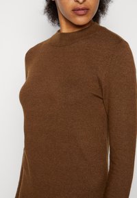 Maglione marrone lavorato a maglia con un colletto alto e maniche lunghe. Il tessuto appare morbido e testurizzato, aderente al corpo.