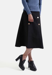 Gonna nera a trapezio con tre grandi bottoni, abbinata a scarpe col tacco nere. La gonna presenta una texture liscia e un design che arriva fino al ginocchio.