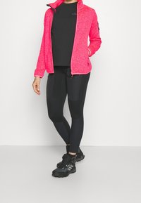 Kobieta nosząca jasnoróżową kurtkę na zamek błyskawiczny, czarne legginsy sportowe z detalami, czarną koszulkę i czarne buty trekkingowe.