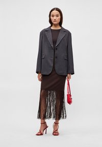 Grijze oversized blazer met twee knopen, gedragen over een bruine jurk met een franjerand. Model draagt een kleine rode handtas. Rode sandalen met hak.