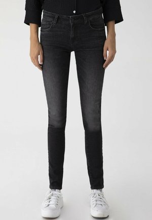 Personne portant un jean skinny noir délavé et des baskets blanches, debout devant un fond clair uni.