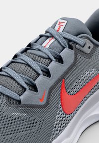 Nike athletic shoe featuring a grey mesh upper, red accents, flat laces, and a textured rubber sole. Lightweight and designed for comfort.