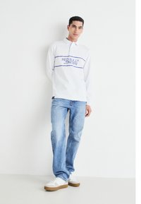 Balta garo piedurkņu polo krekls ar zilu "RED BULL RACING" tekstu, kombinēts ar gaiši ziliem džinsiem un baltām apavām ar brūnu zoli.