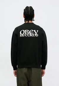 Μαύρο φούτερ με λευκό κείμενο "OBEY RECORDS" σε έντονη τυπογραφία στην πλάτη. Το υλικό φαίνεται μαλακό με χαλαρή εφαρμογή και ριμπ τελείωμα.