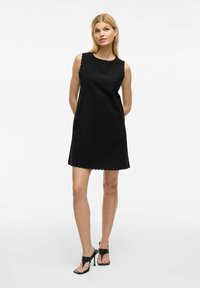 Robe noire sans manches avec un ourlet en scallop et un tissu texturé. Associée à des sandales à talons noirs, se tenant devant un fond uni.