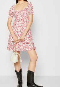 Šaty s květinovým vzorem v bílé a červené barvě, s nabíranými rukávy a čtvercovým výstřihem, doplněné černými kovbojskými botami a malou bílou taškou na šňůrku.