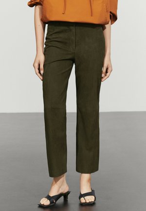 Persona che indossa pantaloni verde scuro a gamba dritta, top arancione e sandali neri con tacco e punta aperta, in piedi su pavimento grigio.
